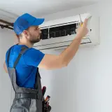 Technik w niebieskiej czapce i ogrodniczkach naprawia klimatyzator.