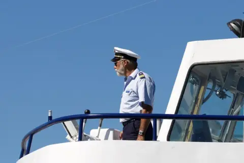 Starszy mężczyzna w białym kapitańskim kapeluszu i okularach przeciwsłonecznych stoi na pokładzie statku.